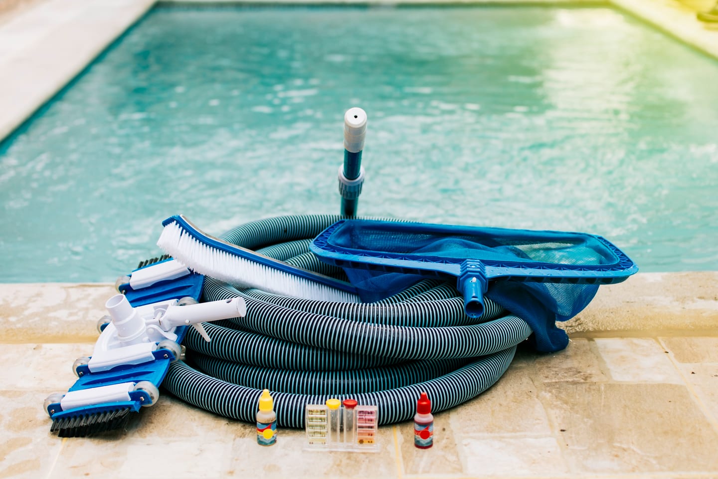 Pool service business starter equipment at poolside — vacuum hose, leaf net, brush, and water test strips for a residential pool route
